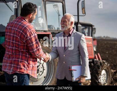 Senior sales representative and mature farmer shaking hands infield in front of tractor Stock Photo