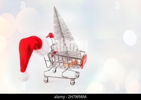 Christmas tree in a supermarket trolley with santa hat on a light background.Simple minimalistic Christmas home decor.Christmas composition.Greeting Stock Photo