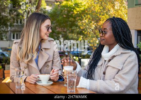 Two young girlfriends enjoy coffee together, sitting at a table, chatting, laughing, and gossiping. Diversity and leisure time concept. Stock Photo