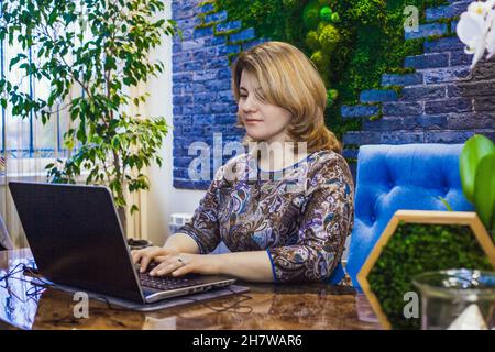 A young woman interior designer is working in her office at a computer. Green office, live plants in an office space, a wall made of stabilized moss, Stock Photo