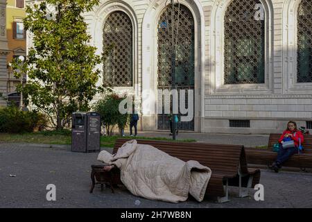 A homeless man in his sleeping bag lies asleep on a bench in front of a church in Bologna, Italy. Stock Photo