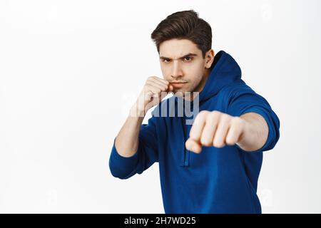 Image of serious man making punch, fighting, wrestle with someone, wearing hoodie, standing over white background Stock Photo