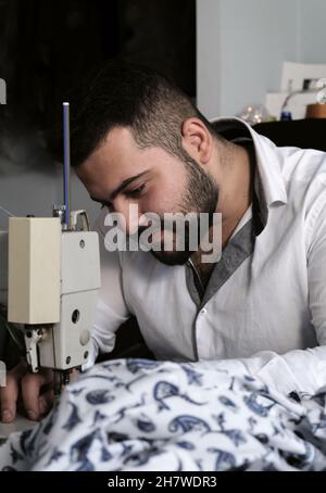 Portrait of Middle eastern male tailor in white shirt working on sewing machine , while makes alterations at client's clothing. Stock Photo