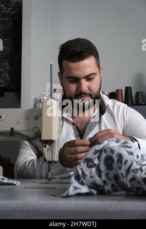 Middle eastern male tailor in white shirt working on sewing machine , while makes alterations at client's clothing. Stock Photo