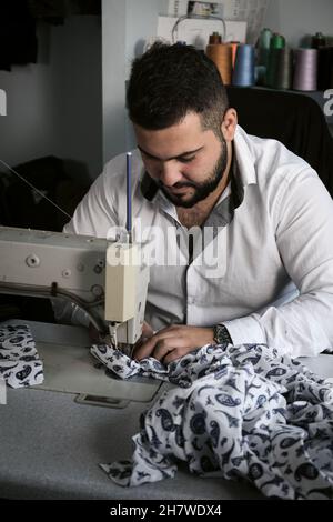 Middle eastern male tailor in white shirt working on sewing machine , while makes alterations at client's clothing. Stock Photo