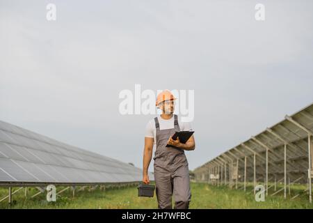 Arabic man in uniform and helmet examining work of solar panels while walking on station. Competent engineer carrying clipboard and instruments in hands. Stock Photo