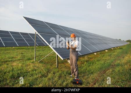 Male arabian engineer in orange helmet and brown overalls checking resistance in solar panels outdoors. Indian man using multimeter while working on station. Stock Photo