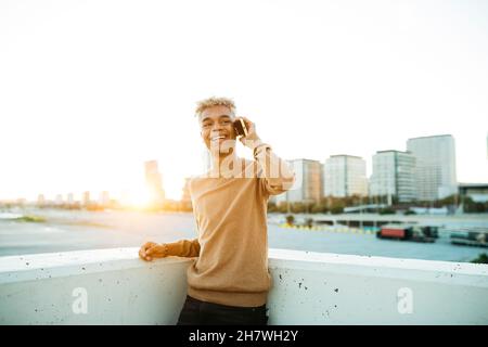 Young latin american male talking and smiling on a yellow smartphone during sunset, on a urban scape Stock Photo