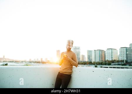 Young latin american male texting and smiling on a yellow smartphone during sunset, on a urban scape Stock Photo