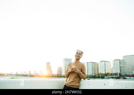 Young latin american male texting and smiling on a yellow smartphone during sunset, on a urban scape Stock Photo