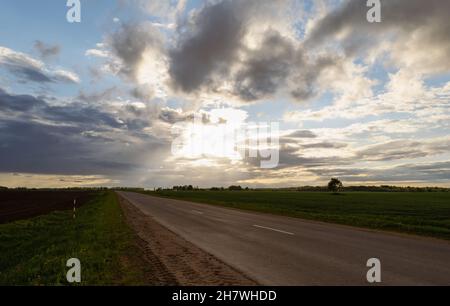 Sunset over the road between fields and meadows. Beautiful clouds. Horizontal photo. Stock Photo