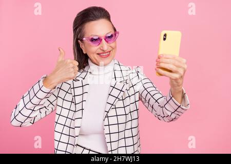 Photo of smiling mature lady in glasses showing thumb-up take selfie with smartphone isolated on pink color background Stock Photo