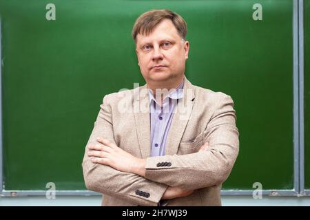 A serious male teacher stands at the green blackboard in the classroom with his arms folded on his chest. Stock Photo
