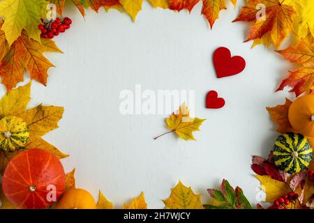 Frame made of autumn leaves, pumpkins and red hearts on a white wooden background. Autumn flat lay with place for text. Horizontal photo. Stock Photo
