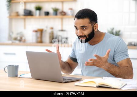 Angry annoyed Indian man, freelancer, manager or designer, sits at a kitchen, emotionally yells at a laptop, got bad news, failed project, lost, gesturing with hands, very irritated and frustrated Stock Photo