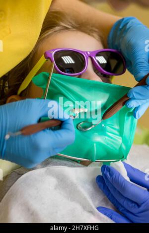 Dentist performing dental filling procedure to preteen girl in pediatric dental clinic. Doctor removing caries using high-speed dental drill. Stock Photo