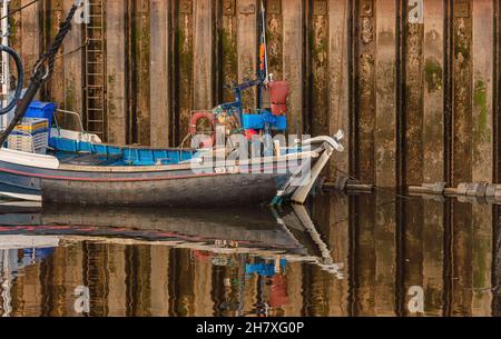 A small fishing boat reflected in the still waters of an harbour. Equipment is stacked at the stern by the tiller.  A ladder runs down form the wharf Stock Photo