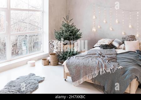 Bedroom with hugo-style bed with gray plaid. natural Christmas tree in pot with christmas lights. Festive decorated bedroom interior with window to fl Stock Photo