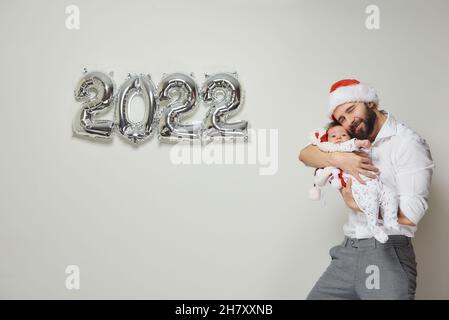 A happy father in a red velvet Santa hat is holding his infant daughter near silver balloons in the shape of 2022. A bearded dad is hugging his little Stock Photo