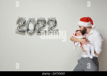 A happy father in a red velvet Santa hat is holding his infant daughter near silver balloons in the shape of 2022. A bearded dad is kissing his little Stock Photo