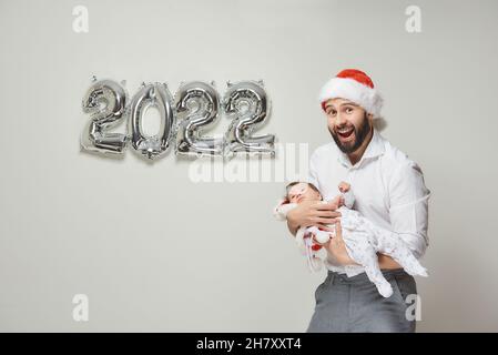 A happy father in a red velvet Santa hat is holding his infant daughter near silver balloons in the shape of 2022. A bearded dad is hugging his little Stock Photo