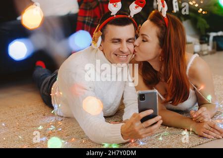 a happy married couple is lying on the floor at home near the Christmas tree and taking pictures of themselves Stock Photo