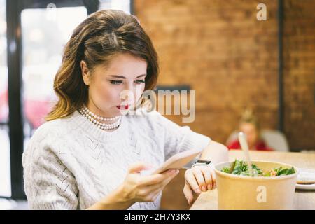 Concentrated Woman looking on her smartphone while having lunch in the cafe. Remote work, social networks scrolling, chatting or making notes Stock Photo
