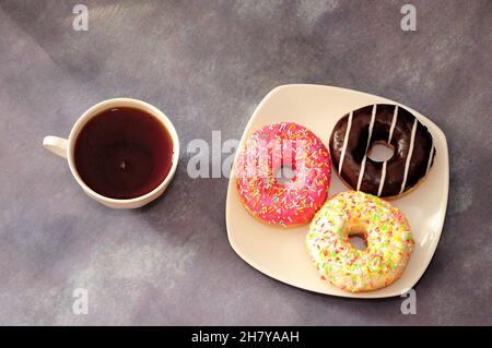 Three donuts in multicolored glaze on a plate and a cup of black tea. Top view, flat lay. Stock Photo
