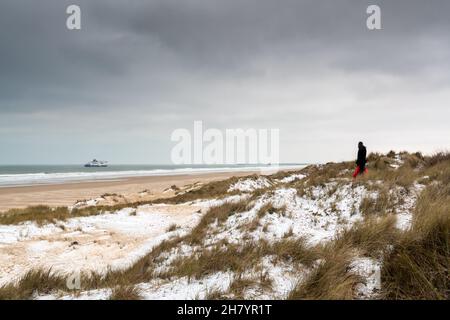 Migrant in the snowy dunes of Sangatte With a simple duvet as luggage, in winter. He's watching a ferry entering the port of Calais from England Stock Photo