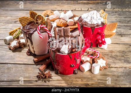 Christmas styled overloaded hot chocolate mugs with various toppings – chocolate slices, spices, sweets, cookies, candy and gingerbreads, wooden cozy Stock Photo