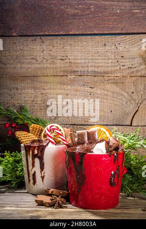 Christmas styled overloaded hot chocolate mugs with various toppings – chocolate slices, spices, sweets, cookies, candy and gingerbreads, wooden cozy Stock Photo