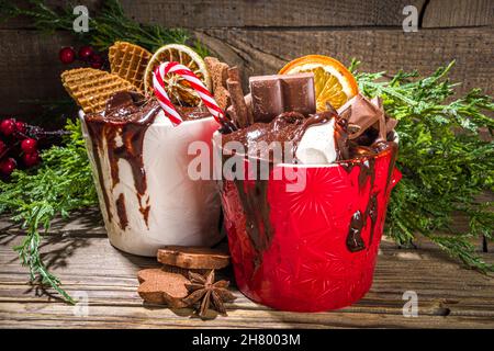 Christmas styled overloaded hot chocolate mugs with various toppings – chocolate slices, spices, sweets, cookies, candy and gingerbreads, wooden cozy Stock Photo