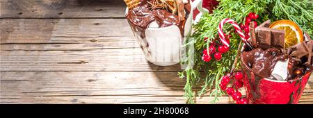 Christmas styled overloaded hot chocolate mugs with various toppings – chocolate slices, spices, sweets, cookies, candy and gingerbreads, wooden cozy Stock Photo