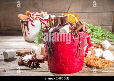 Christmas styled overloaded hot chocolate mugs with various toppings – chocolate slices, spices, sweets, cookies, candy and gingerbreads, wooden cozy Stock Photo