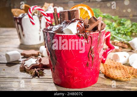 Christmas styled overloaded hot chocolate mugs with various toppings – chocolate slices, spices, sweets, cookies, candy and gingerbreads, wooden cozy Stock Photo