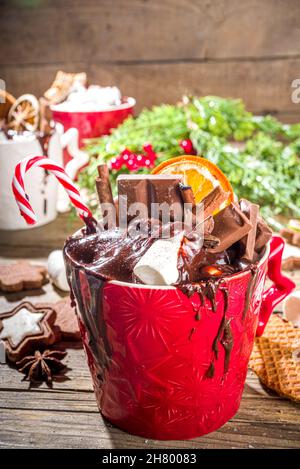 Christmas styled overloaded hot chocolate mugs with various toppings – chocolate slices, spices, sweets, cookies, candy and gingerbreads, wooden cozy Stock Photo