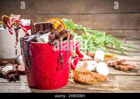 Christmas styled overloaded hot chocolate mugs with various toppings – chocolate slices, spices, sweets, cookies, candy and gingerbreads, wooden cozy Stock Photo