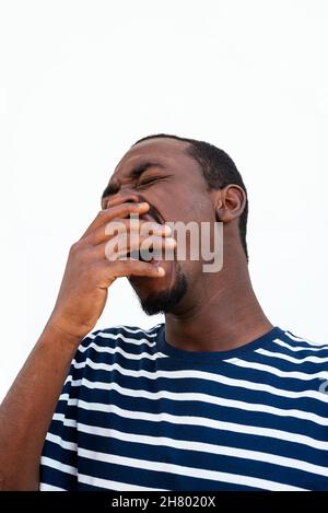 Tired bearded African American male in stylish striped shirt covering mouth with hand while yawning on white background in city Stock Photo
