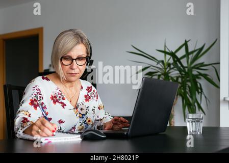 Concentrated female remote worker in eyeglasses sitting at table and writing notes in planner while doing job on netbook at home Stock Photo