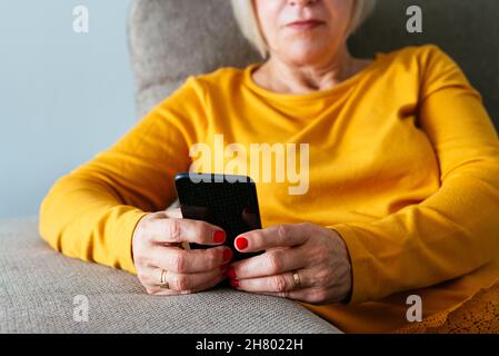 Anonymous middle aged female sitting on sofa and browsing mobile phone at home Stock Photo