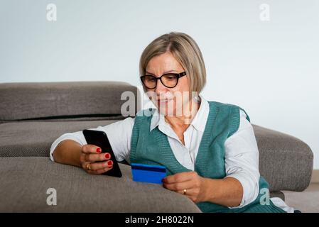 Middle age female sitting on sofa and paying for online purchases with credit card and cellphone Stock Photo