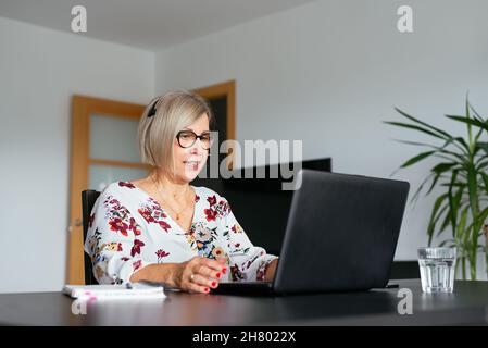 Concentrated middle age female remote specialist in eyeglasses and headset working on netbook during telework at home Stock Photo