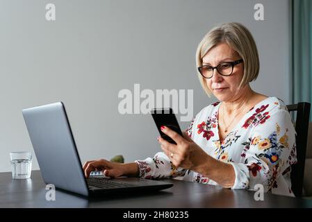 Middle age female remote worker in eyeglasses working on laptop while using mobile phone at table Stock Photo