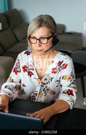 Concentrated middle age female remote specialist in eyeglasses and headset working on netbook during telework at home Stock Photo