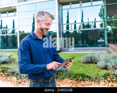 Focused bearded male in shirt and jeans scrolling tablet while dealing with work issues remotely in park near modern office building Stock Photo