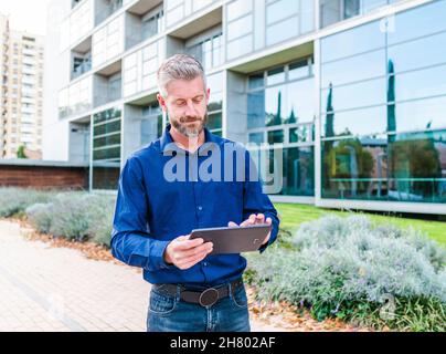 Focused bearded male in shirt and jeans scrolling tablet while dealing with work issues remotely in park near modern office building Stock Photo