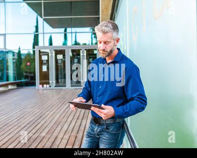 Focused bearded male worker browsing tablet while standing near modern business center and working on project Stock Photo