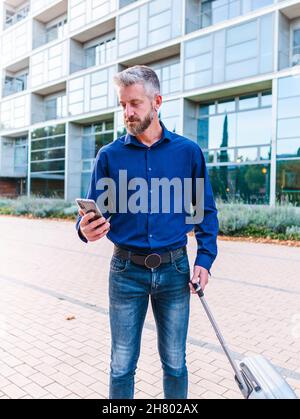 https://l450v.alamy.com/450v/2h802ax/serious-gray-haired-bearded-male-executive-worker-with-suitcase-using-cellphone-near-modern-tower-during-business-trip-2h802ax.jpg