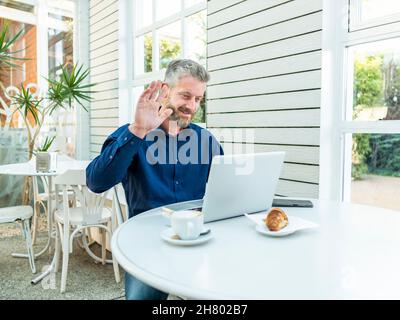 Smiling male entrepreneur waving hand to camera of netbook during online video chat at table with smartphone and coffee with croissant in outdoor cafe Stock Photo