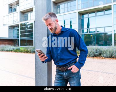 Adult male with gray hair and beard standing with hand in pocket of jeans near pillar and browsing mobile phone Stock Photo